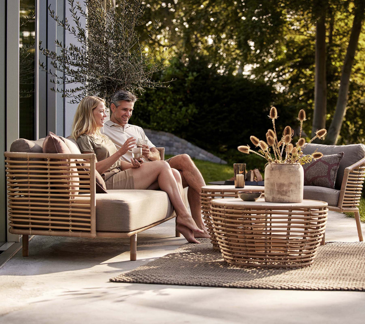A couple enjoys drinks on a Sense sofa and lounge chair, accompanied by a stylish basket coffee table and decorative vase.