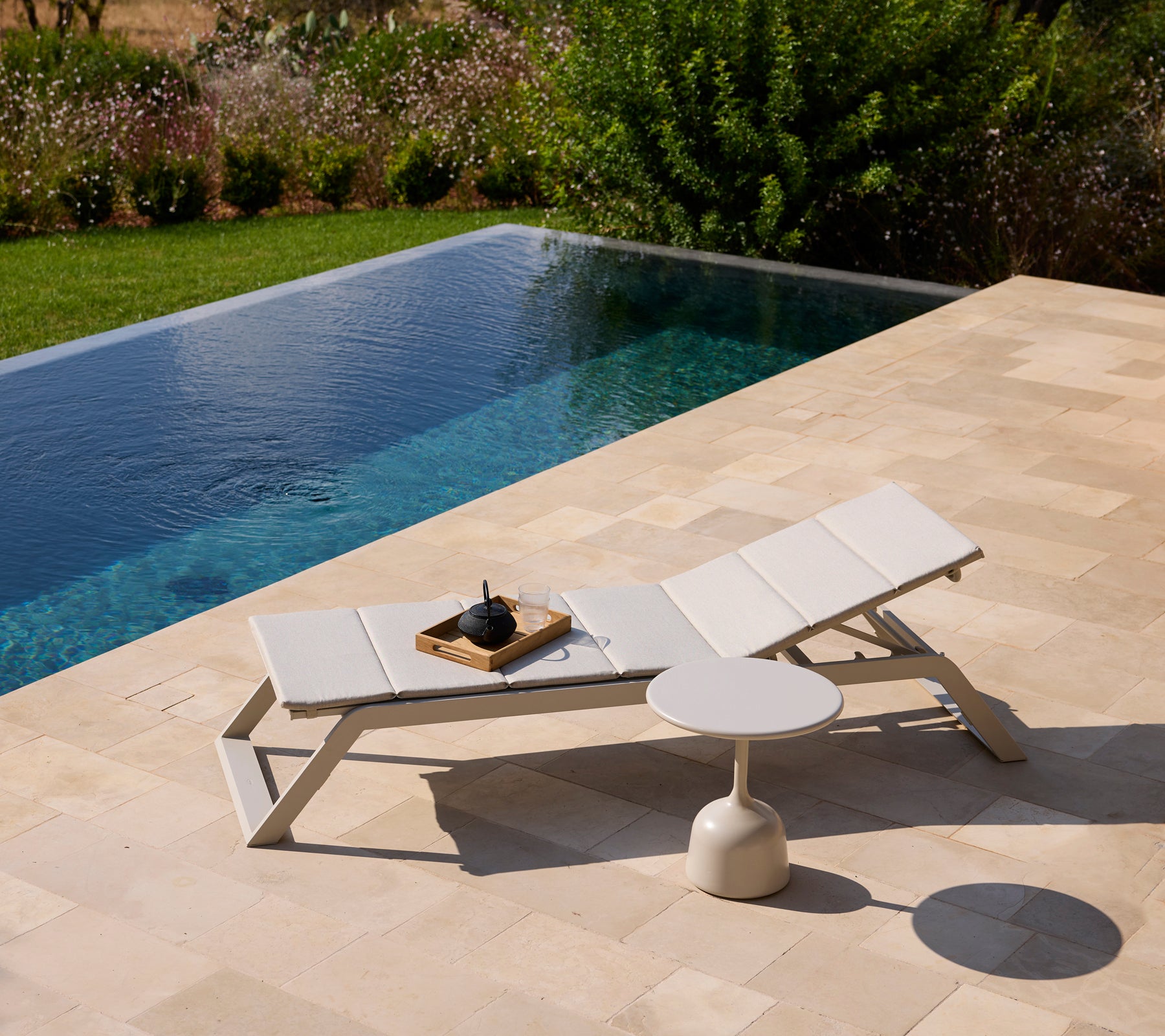 Modern white lounge chair and side table by a pool surrounded by greenery.