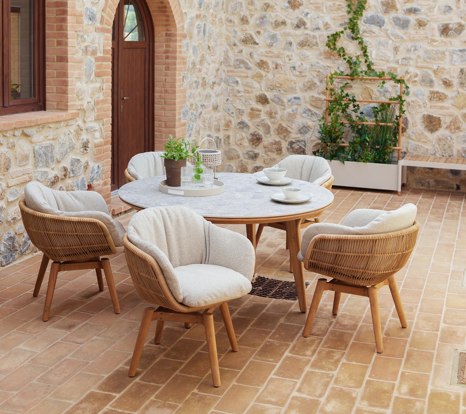 A stylish dining setup featuring a round table surrounded by swivel chairs with teak legs and comfortable cushions, complemented by greenery.