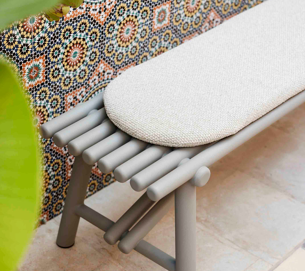 Taupe aluminium bench with stick-like legs and a rounded cushion, set against a colorful patterned backdrop.