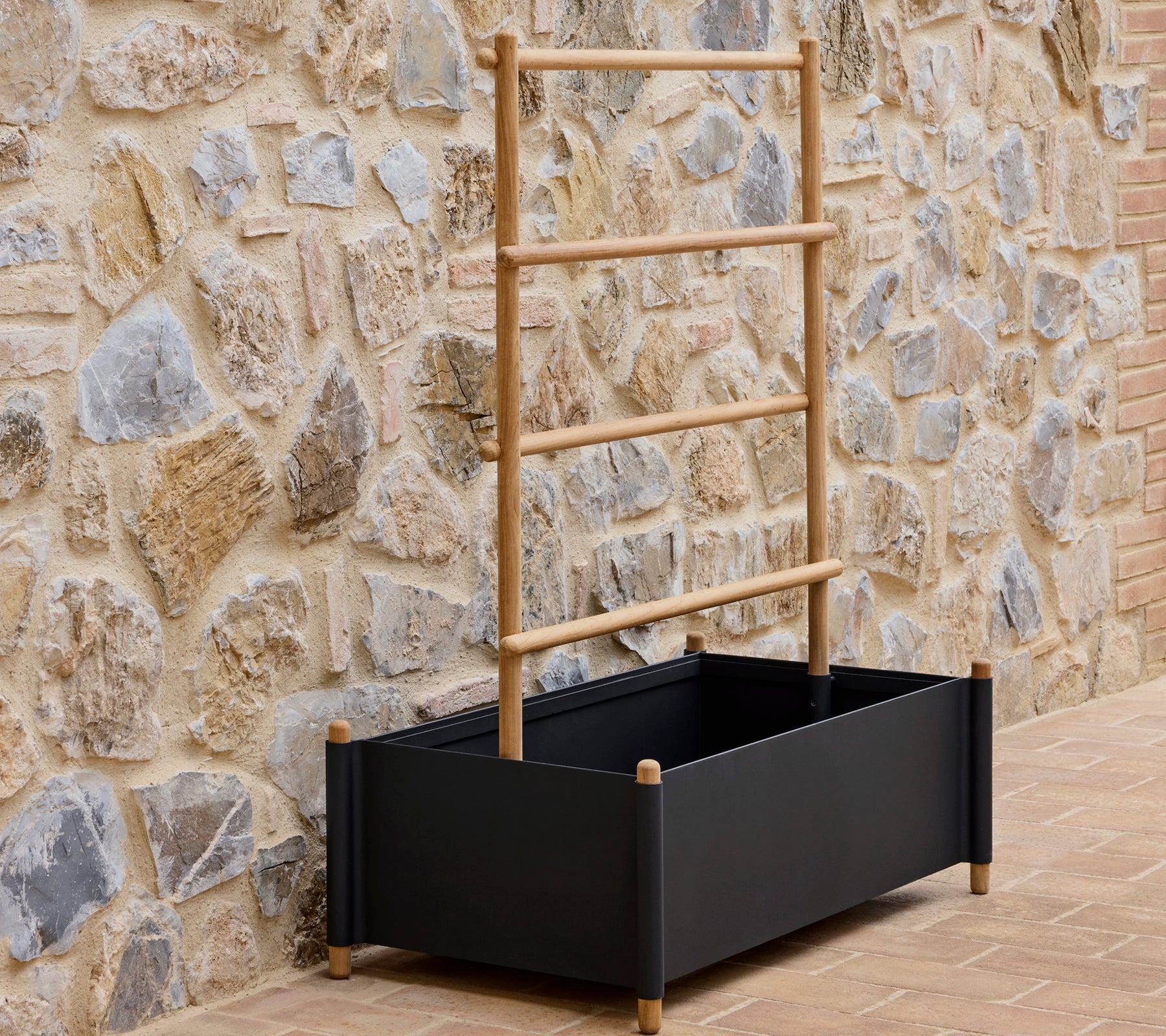 Black aluminum planter with wooden support sticks, placed against a textured stone wall.