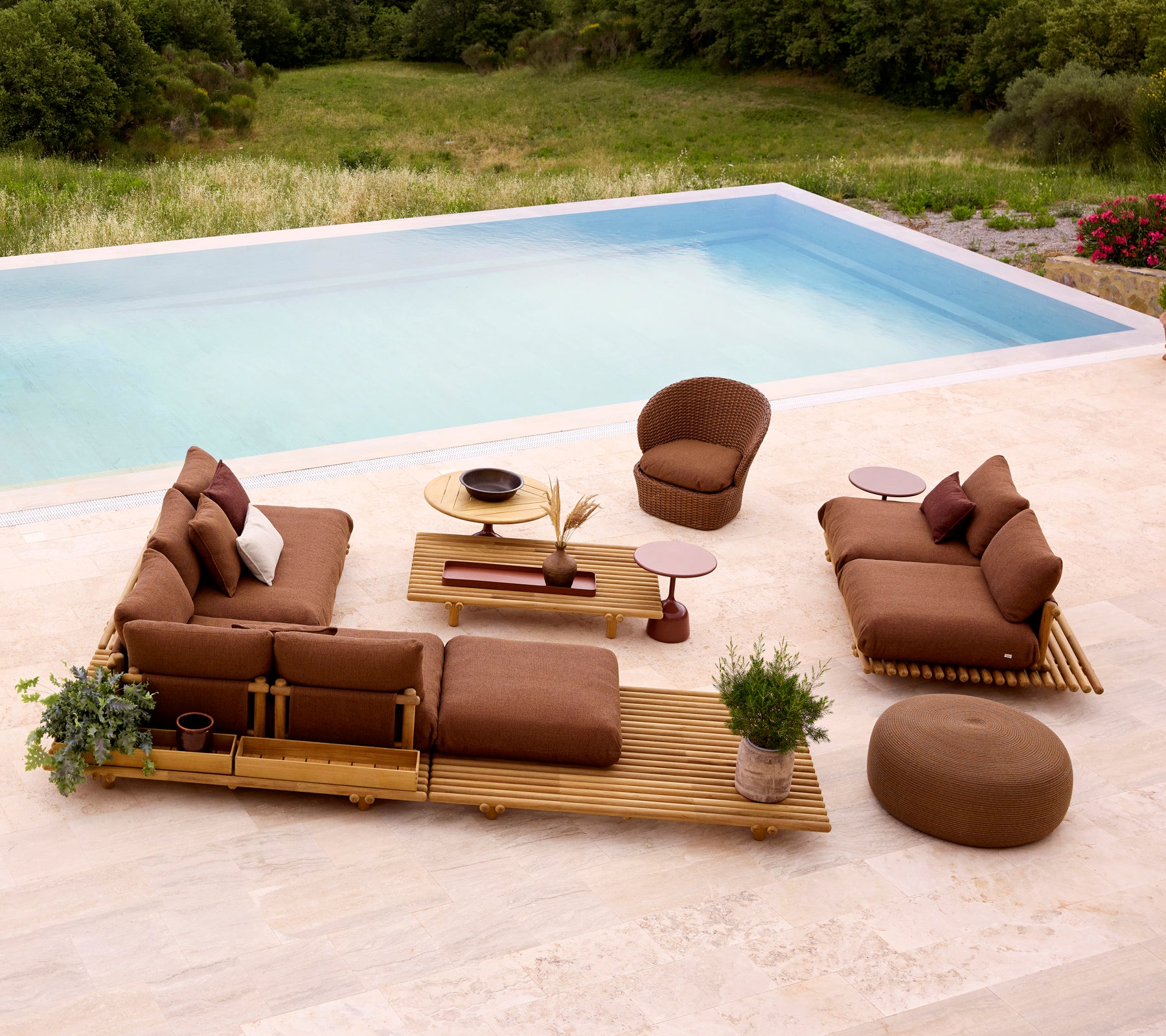 Stylish lounge setting featuring teak furniture, plush cushions, and a round footstool, complemented by greenery and a table.