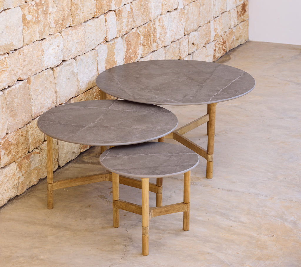 Three modern grey coffee tables with light wooden bases against a stone wall.