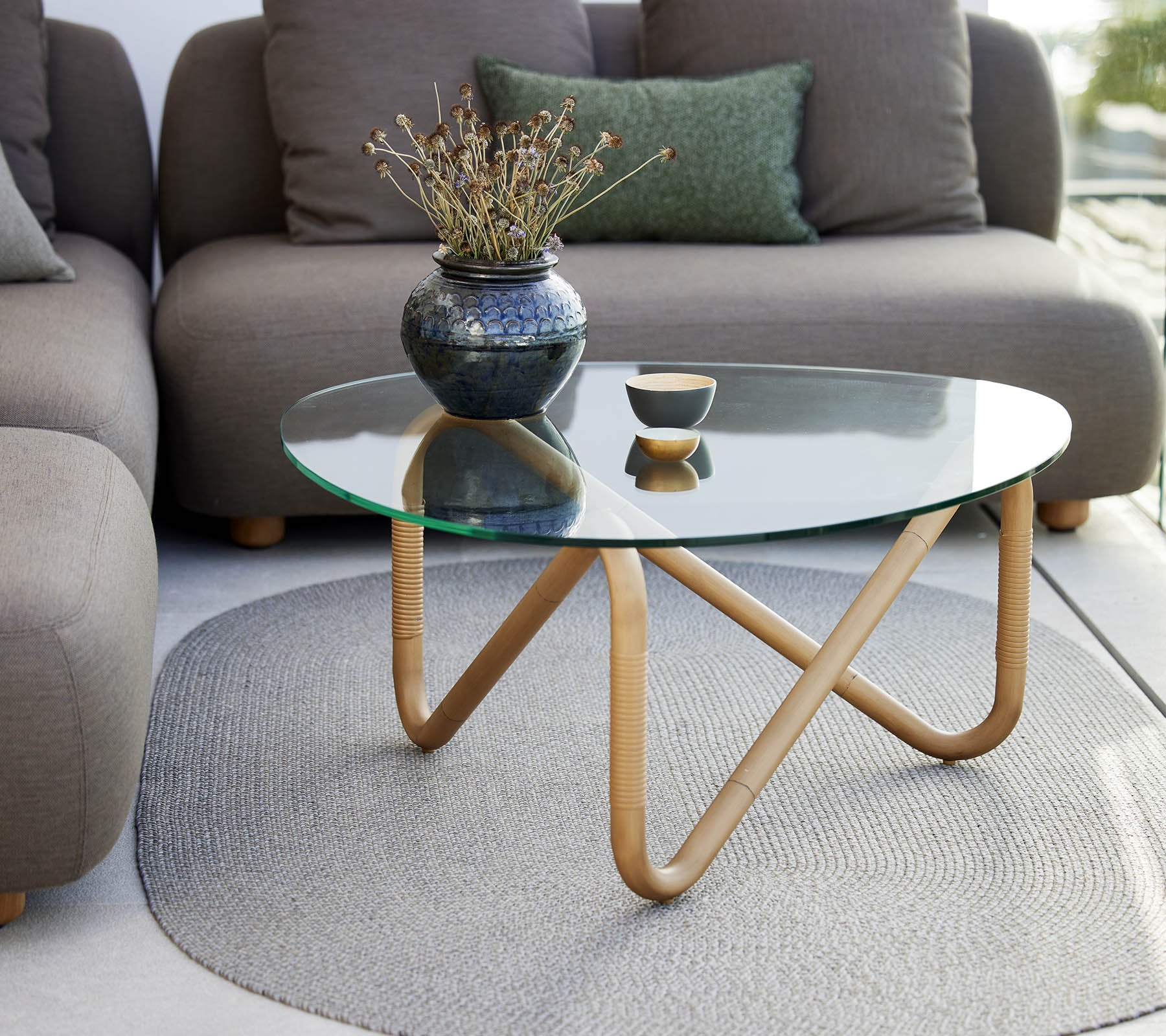 Wave coffee table with a glass top and wooden base, accented by a vase and a small bowl, surrounded by soft, neutral-colored seating.