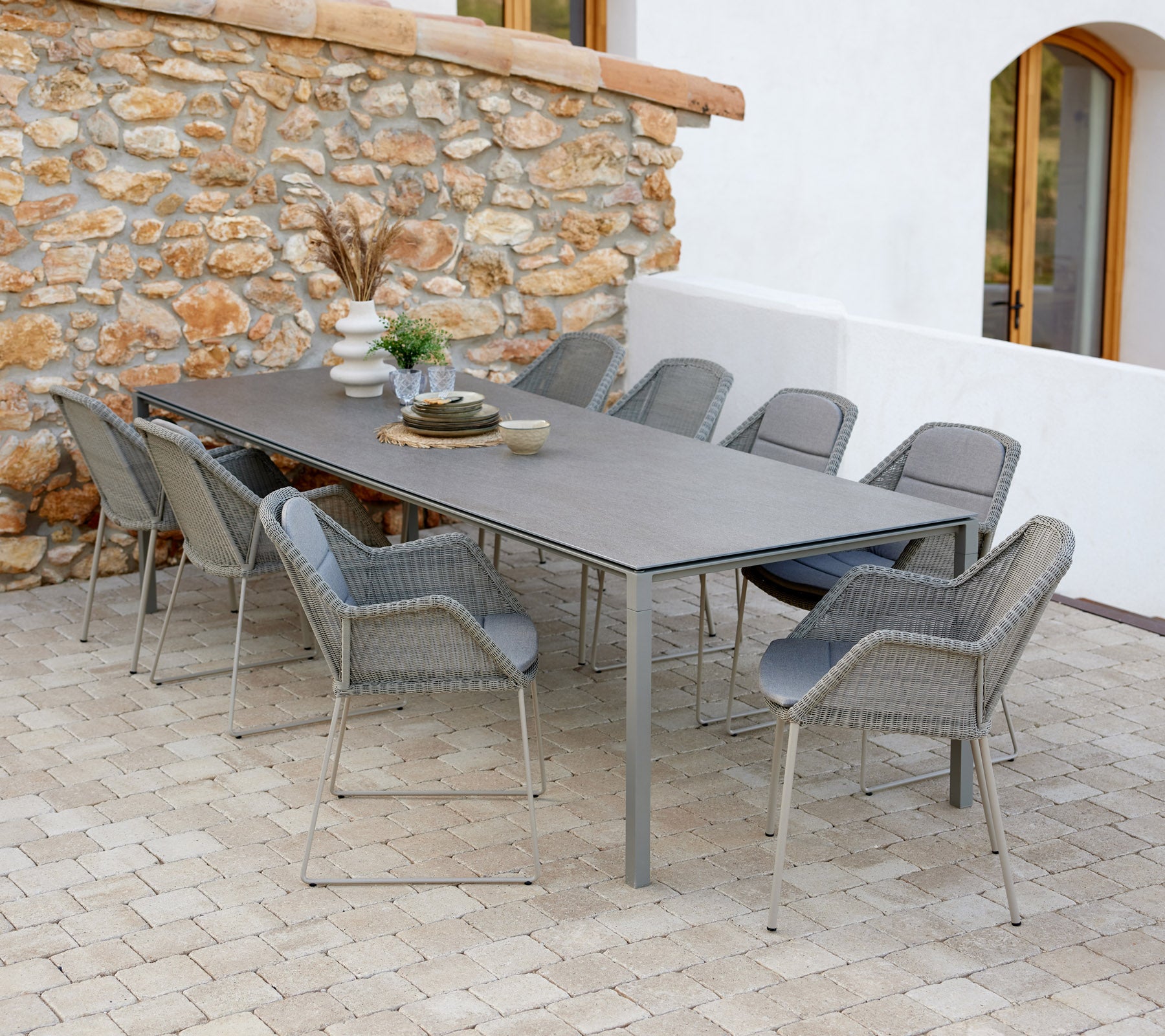 Modern dining table with taupe chairs, set with decorative items and a touch of greenery against a stone wall backdrop.