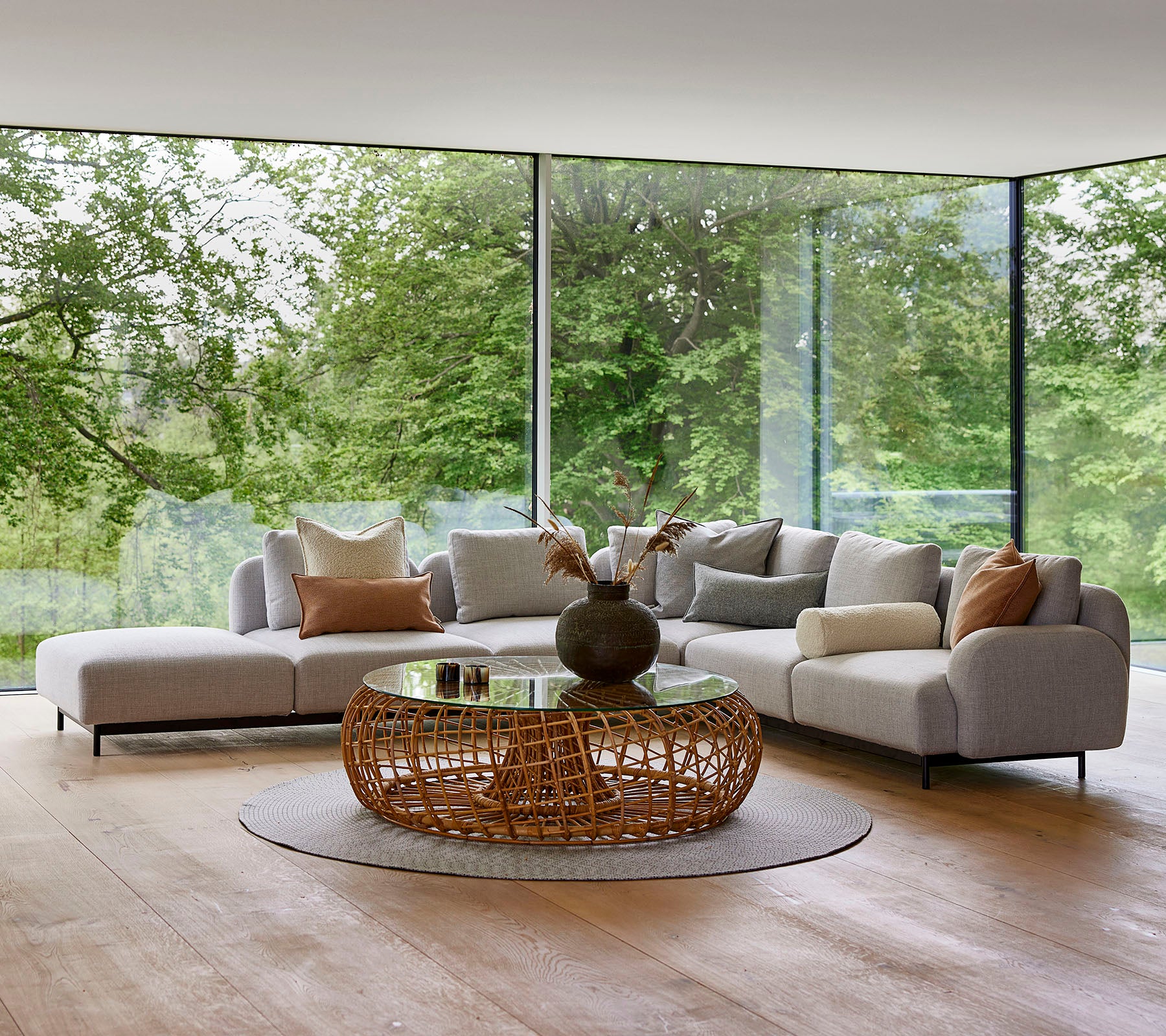 Modern lounge area featuring a modular sofa with decorative pillows, a round woven coffee table, and large glass windows showcasing greenery.