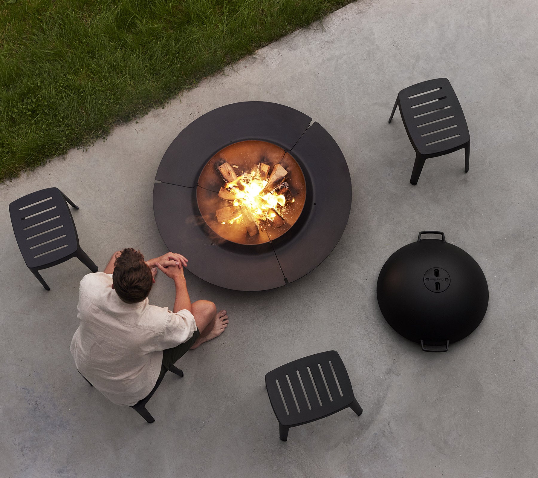 A person sits near a circular fire pit surrounded by four black chairs, enjoying a cozy fire with logs and a round lid nearby.